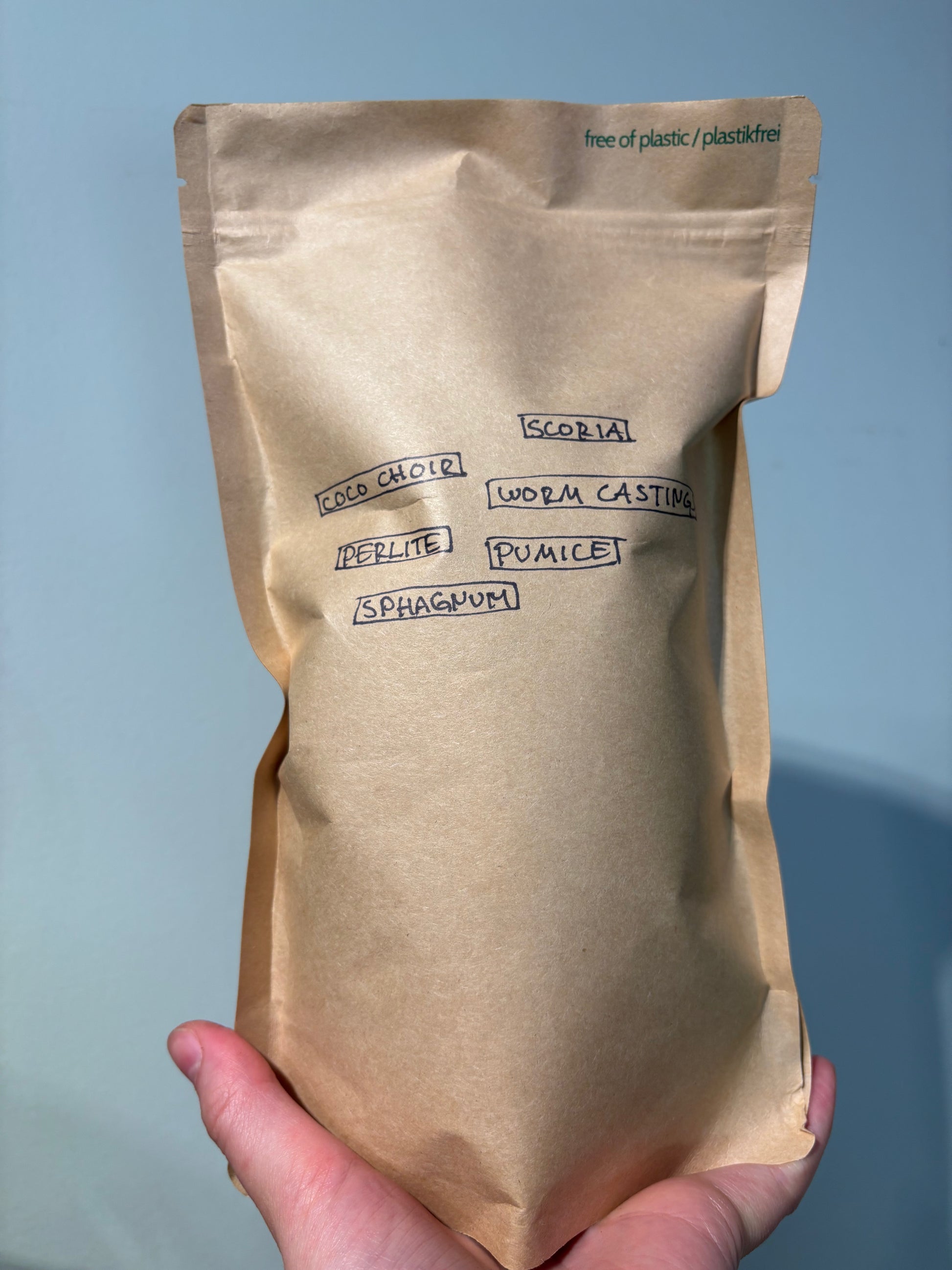 Hand holding labeled kraft paper bag of Home made Aroid Soil Mix listing ingredients like coco coir, perlite, pumice, and worm castings.